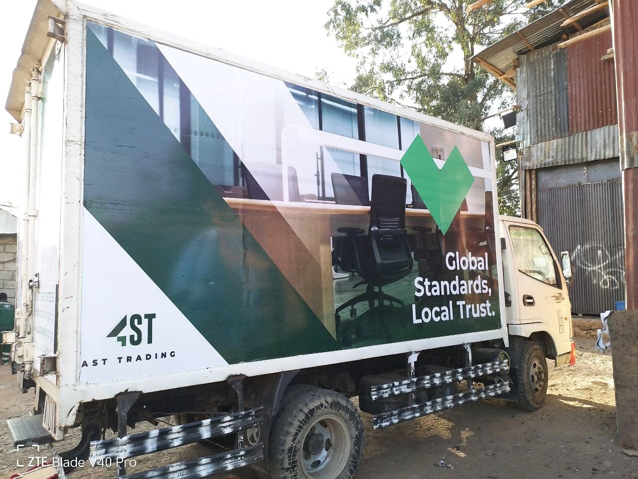 AST Trading box truck printed with a green diagonal brand device, an office interior photograph and the line "Global Standards, Local Trust."