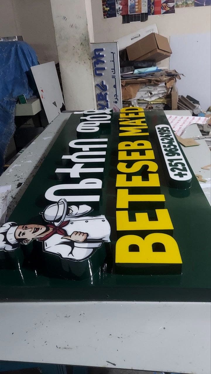Deep green restaurant sign panel with raised white Amharic and yellow Latin lettering and a cut-out chef illustration, photographed flat on the workshop bench