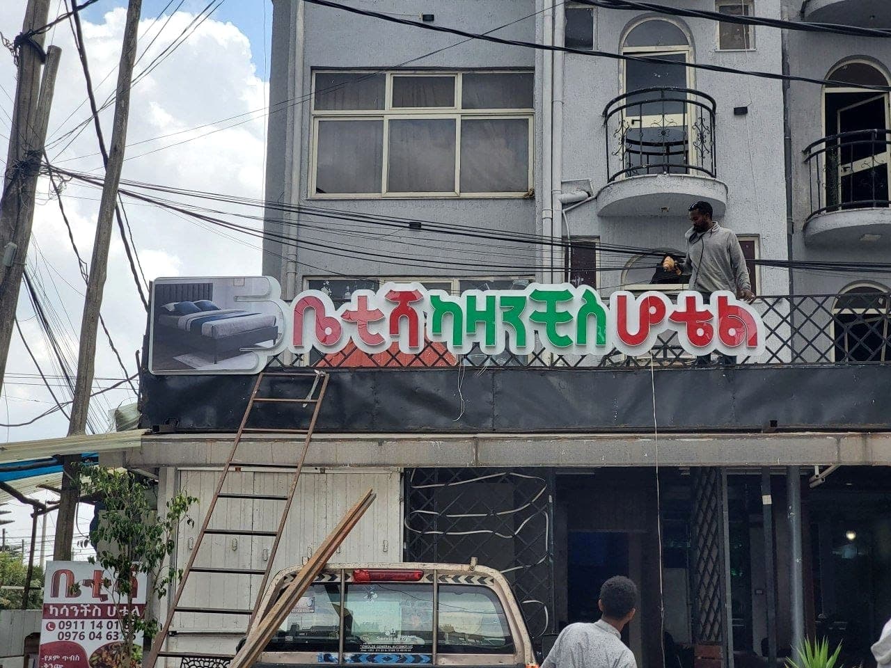 Red and green illuminated Amharic lettering for Betesh Casanchis Hotel being fixed to a balcony fascia, with a fitter on the balcony above