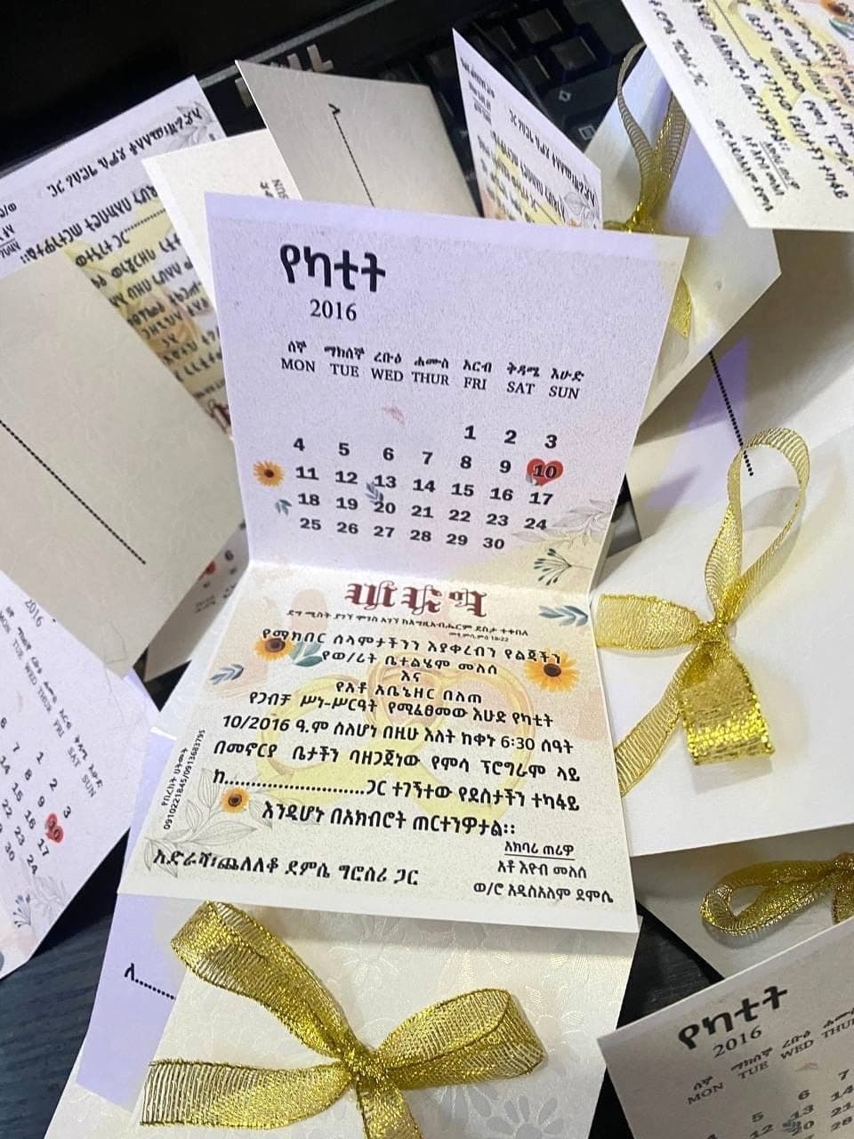 Folded invitation cards with a printed Amharic calendar inside, tied with gold ribbon