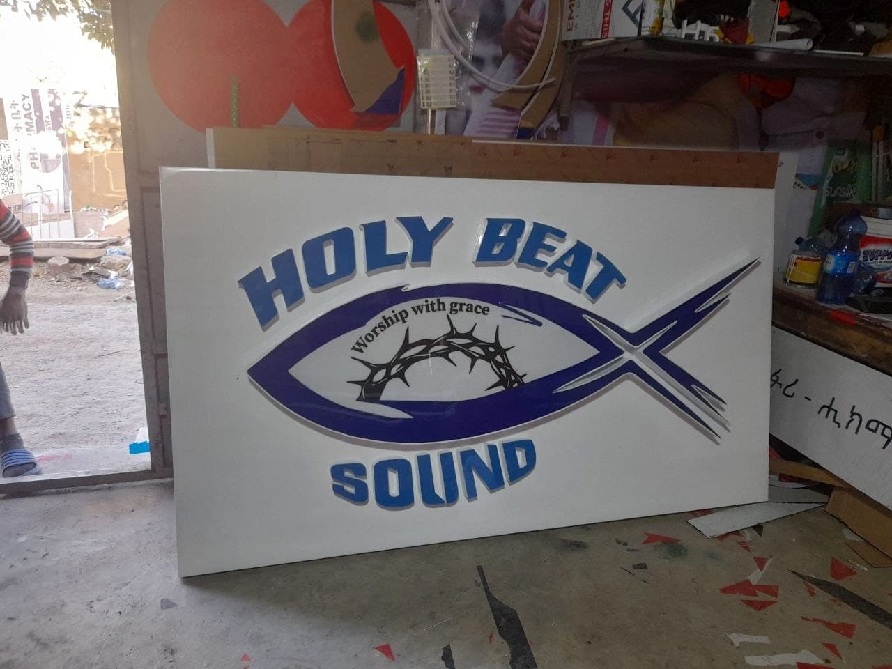 White sign panel with raised blue Holy Beat Sound lettering around an ichthys and crown-of-thorns motif, standing in the workshop doorway