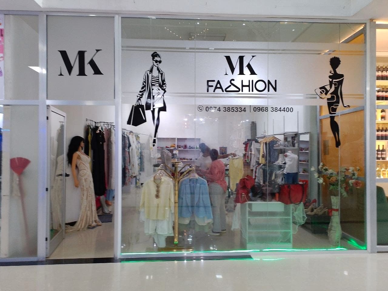 Glass shopfront with the MK Fashion wordmark and two cut-vinyl fashion illustrations applied, the shop lit and stocked behind