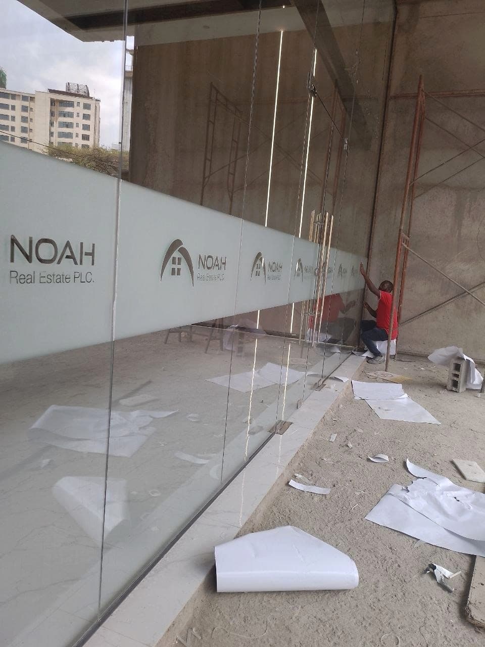 Frosted manifestation banding carrying the repeated Noah Real Estate logo being applied along a full-height glazed frontage, with the fitter working along the run