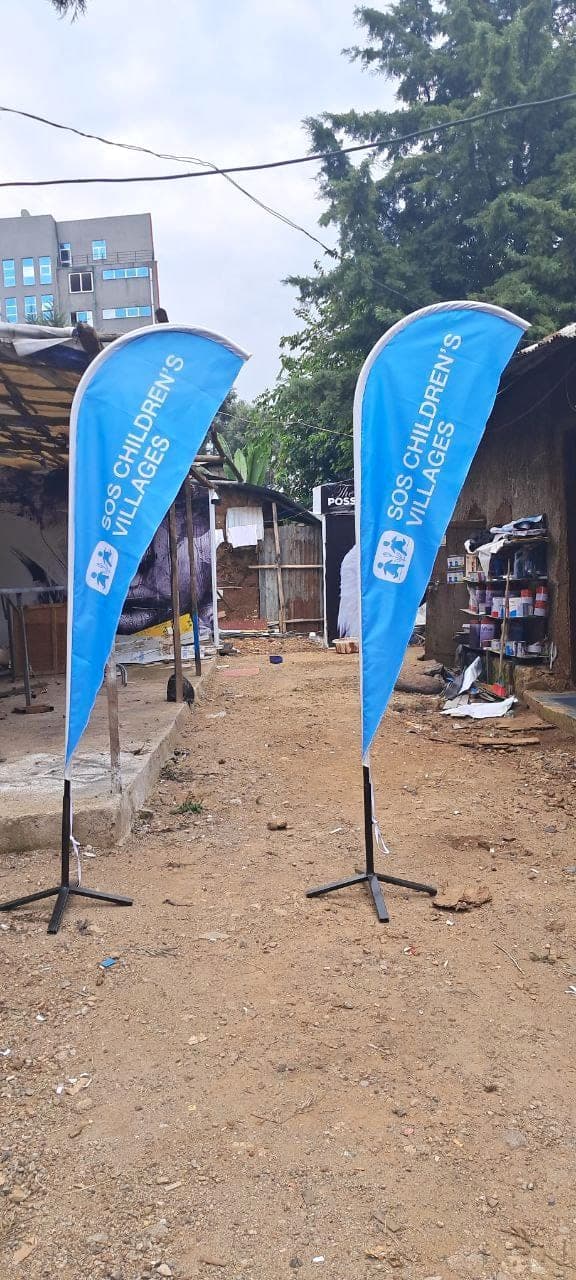 Pair of blue SOS Children's Villages teardrop flags on cross bases, standing outside the workshop