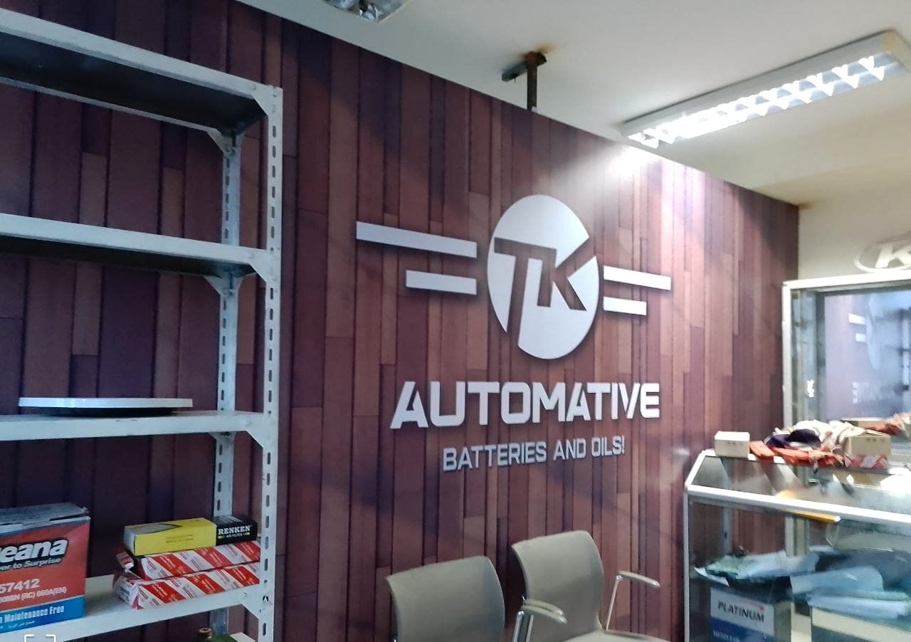 Brushed metal TK Automative logo and lettering reading "Batteries and Oils" mounted on a dark timber-clad showroom wall