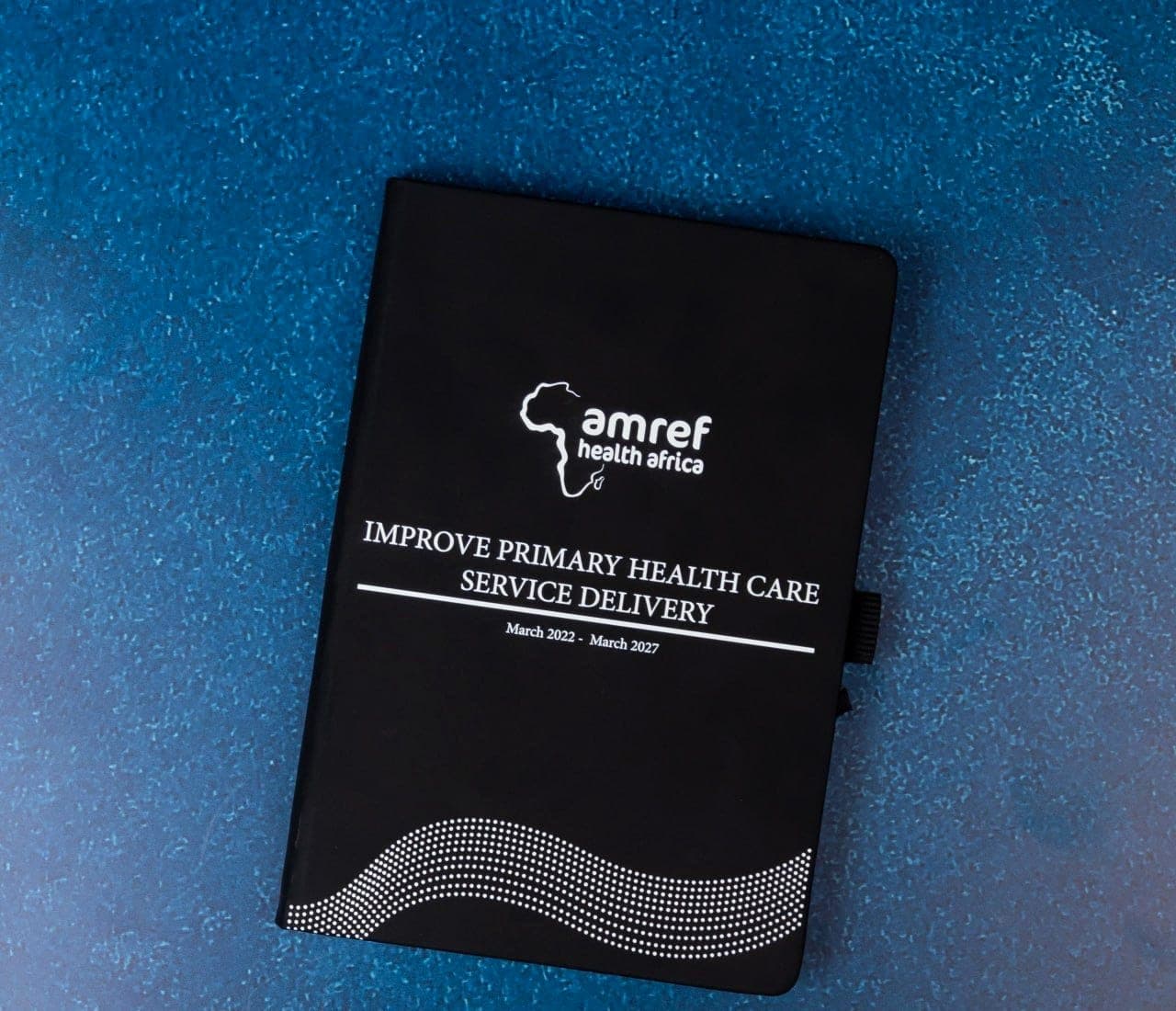 Black hardback notebook printed with the Amref Health Africa logo and a primary health care programme title, on a blue background