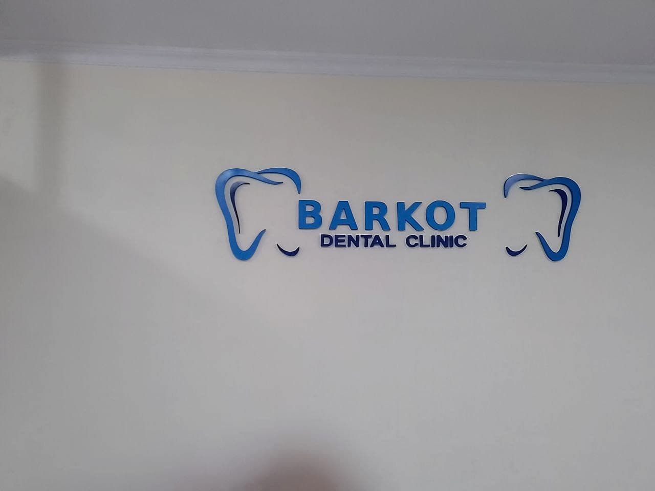 Blue dimensional letters reading Barkot Dental Clinic flanked by two cut tooth motifs on a pale reception wall
