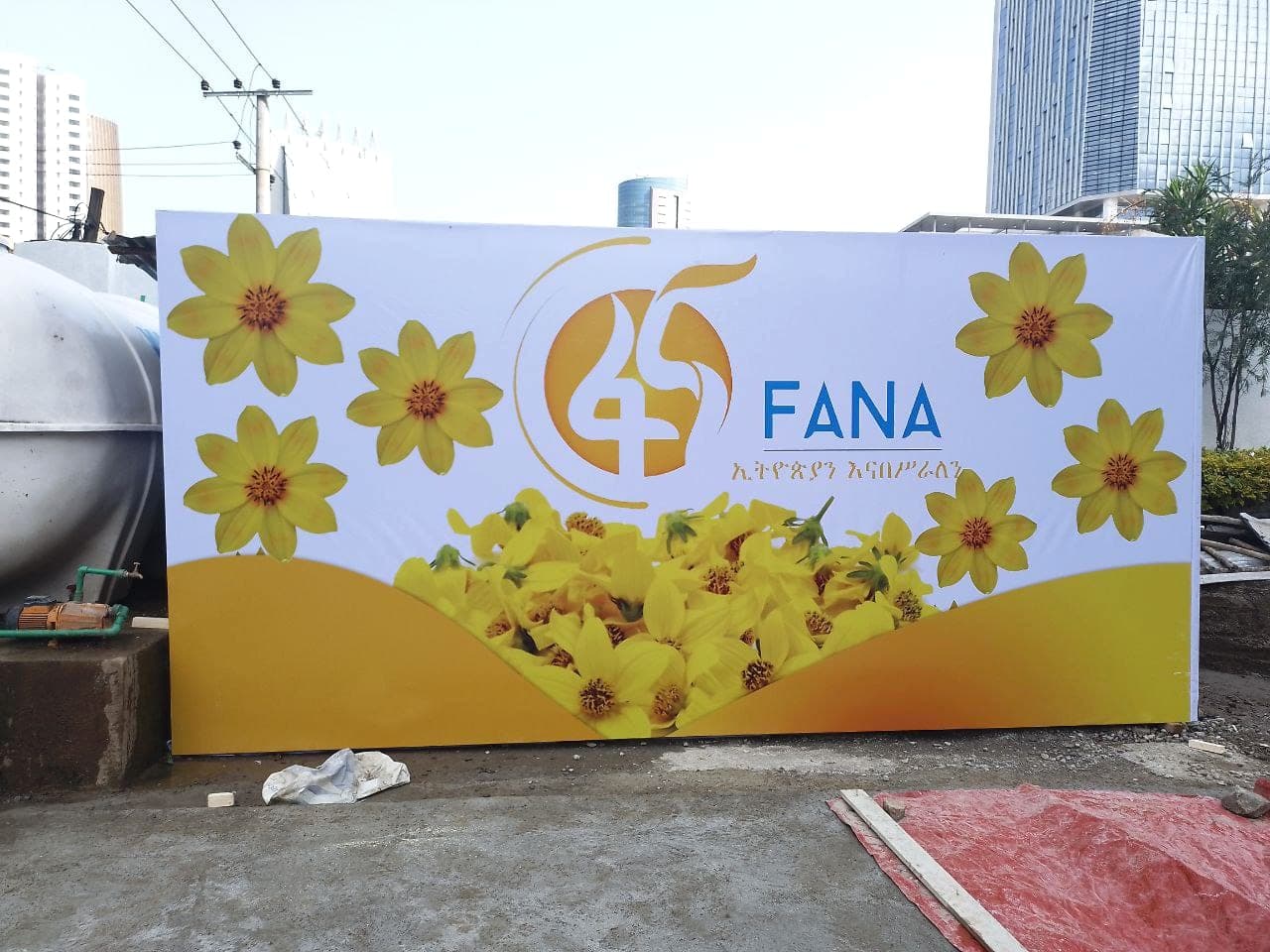 Wide Fana 45th anniversary hoarding panel printed with yellow meskel flowers, standing in the yard before installation