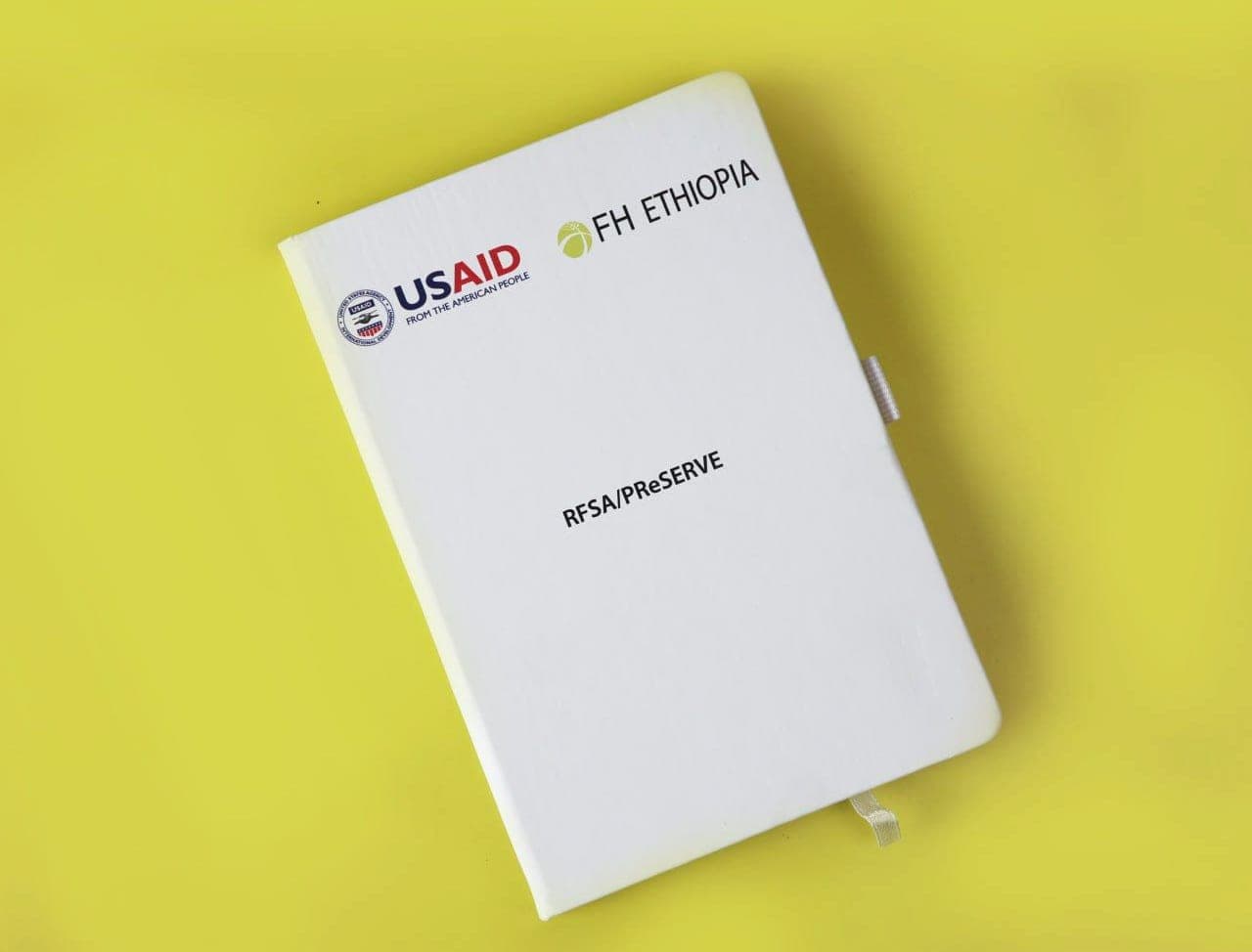 White hardback notebook printed with the USAID and FH Ethiopia logos and the RFSA/PReSERVE programme name, on a yellow background